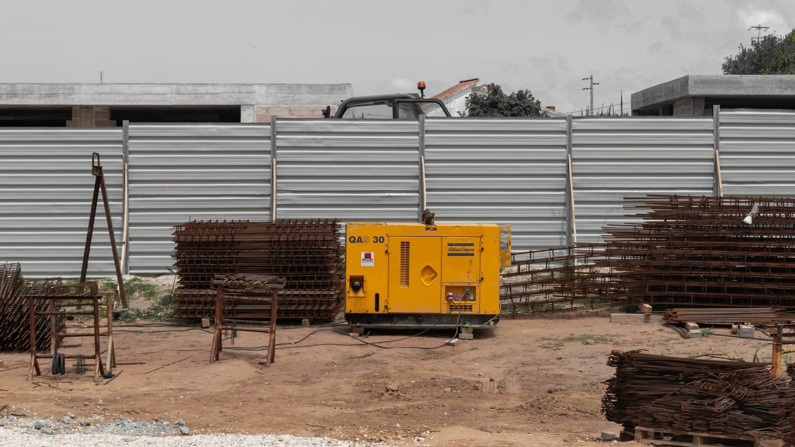 A yellow diesel generator on bare ground beside stacks of steel reinforcing bars